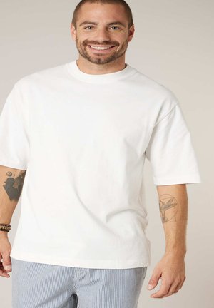Homme souriant portant un t-shirt blanc uni surdimensionné et un pantalon rayé, avec des tatouages visibles sur les deux avant-bras et des bracelets de perles au poignet gauche.