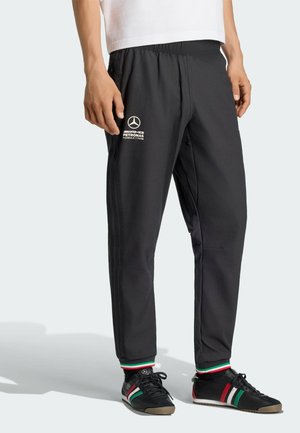 Männliches Model trägt schwarze Mercedes AMG Petronas Trainingshose mit elastischem Bund sowie Socken und Schuhe mit italienischem Flaggenmuster.