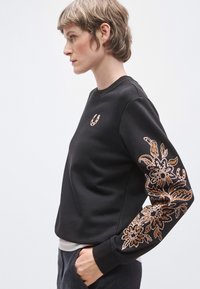 Μαύρο φούτερ με μακριά μανίκια, με περίπλοκη κεντητική με σχέδια λουλουδιών σε χρυσό και μπεζ στο αριστερό μανίκι και ένα λογότυπο στο στήθος.