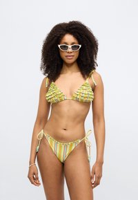 Bikini komplekts ar dzeltenas krāsas volāniem un daudzkrāsainām svītrām; sānus un plecu lencītes iespējams sasiet, tām ir dekoratīvi pērlīšu rotājumi.