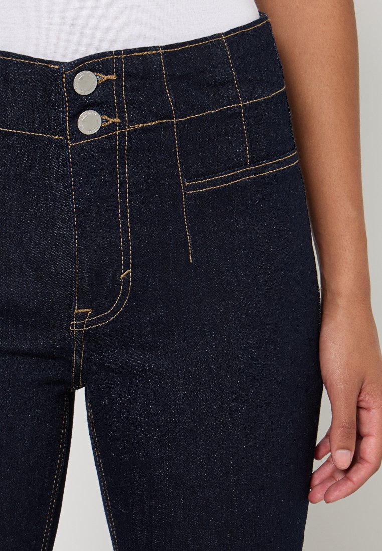 Jeans en denim bleu foncé avec une taille haute, fermeture à double bouton et coutures dorées. Poches avant visibles avec une surface lisse.