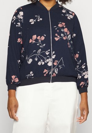 Vrouw draagt een marineblauwe bomberjack met rits, voorzien van roze en witte bloemenpatronen, en witte broek, tegen een effen achtergrond.