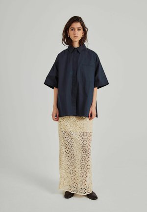 Marineblauw kortekoffieshirt met kraag en zak, gecombineerd met een beige gebreide maxi-jurk van kant met hexagonale en bloemmotieven.