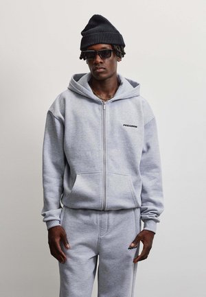 Junger Mann trägt einen grauen Kapuzenpullover mit Reißverschluss und passende Jogginghose, schwarze Mütze, dunkle Sonnenbrille und eine goldene Halskette vor einem schlichten Hintergrund.