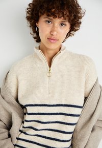 Knit sweater with a quarter-zip collar in beige with vertical stripes. Soft texture and gold hardware accent. Warm, casual design.