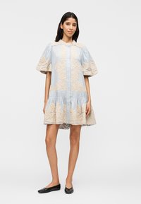 Mujer de pie con un vestido azul claro abotonado, mangas abullonadas y bordado floral beige, combinado con zapatos planos negros.