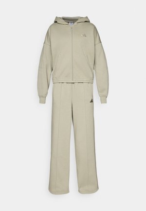 Felpa zip in beige chiaro con cappuccio, maniche ampie e logo ricamato; abbinata a pantaloni a gamba larga con vita elasticizzata.