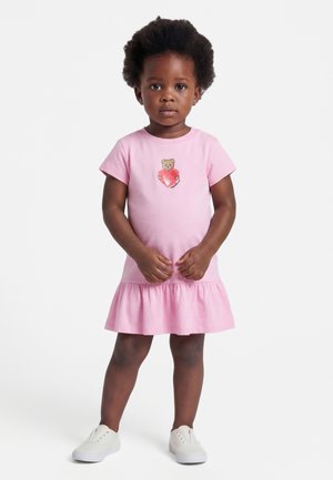 Μικρό παιδί που στέκεται φορώντας ένα ροζ φόρεμα με κοντά μανίκια, διακοσμημένο με σχεδιασμό αρκουδάκι και καρδιά, συνδυασμένο με λευκά αθλητικά παπούτσια.