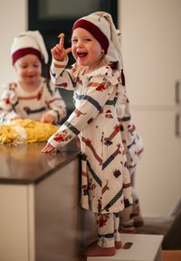 Lapset, jotka pukeutuvat vaaleanbeigeihin pyjamapaketeihin, joissa on koira- ja lahjatulostuksia, yhdistettynä punaisiin ja valkoisiin hattuihin, leikkivät keittiötasolla.