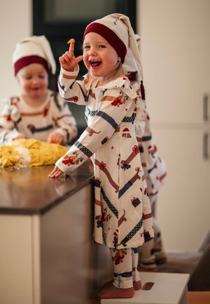 Lapset, jotka pukeutuvat vaaleanbeigeihin pyjamapaketeihin, joissa on koira- ja lahjatulostuksia, yhdistettynä punaisiin ja valkoisiin hattuihin, leikkivät keittiötasolla.
