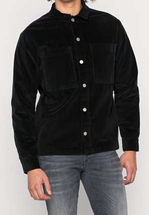 Uomo con camicia nera di velluto a coste con tasche sul petto e jeans grigio chiaro, in piedi davanti a uno sfondo semplice.