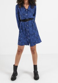 Langärmliges blaues Kleid mit gepunktetem Muster, Knöpfen vorne und einem schwarzen Gürtel in der Taille. Kombiniert mit schwarzen Knöchelschuhen.