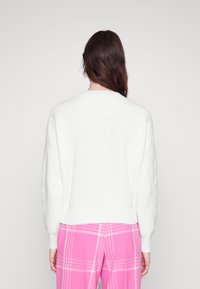 Maglione bianco lavorato a maglia con texture a coste e scollo rotondo, abbinato a pantaloni a quadri rosa, visto da dietro.