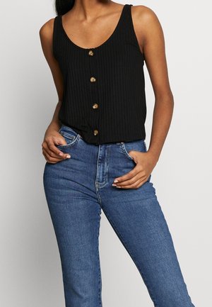 Kvinde iført sort ribbet, knaplukket top uden ærmer og højtaljede blå jeans, stående med hænderne i lommerne mod en ensfarvet baggrund.