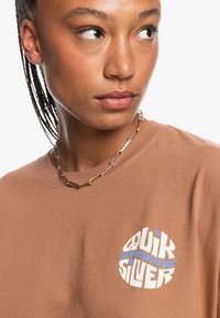 Bruine katoenen t-shirt met een ronde blauwe en witte graphic met de tekst "Quiksilver". Zachte textuur met een ronde hals en korte mouwen.