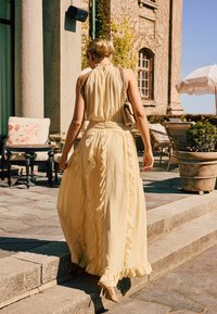 Femme en robe jaune pâle à volants et talons montant des marches en pierre devant un bâtiment avec une terrasse extérieure et des plantes.