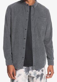 Szary kordurojowy koszula zapinana na guziki z kołnierzykiem, kieszenią na piersi oraz czarnymi guzikami, noszona na czarnym t-shircie, w zestawieniu z spodniami w tie-dye.