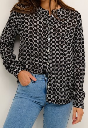 Frau trägt schwarzes Hemd mit weißen geometrischen Mustern und hellblauen Jeans, mit einer Hand in der Hosentasche.