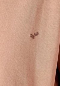 Tejido de color rosa claro con una textura suave que presenta un pequeño detalle de punto de cruz en marrón oscuro. Diseño simple sin patrones adicionales.