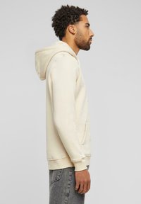 Kremfarget hettegenser med lommer foran, justerbar snor i hetta, ribbede mansjetter og løs passform, kombinert med grå slitte jeans.
