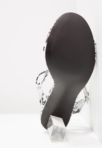 Sandale à talon à motifs noir et blanc avec une semelle noire texturée et un talon bloc transparent, dotée de fines lanières et d'une quincaillerie minimale.