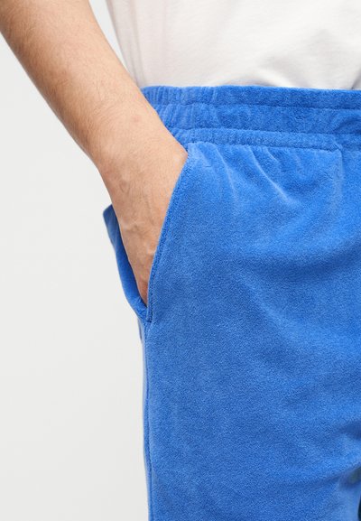 Χέρι που ακουμπά μέσα στην τσέπη φωτεινών μπλε βερμούδων από τερι ύφασμα με ελαστική μέση, σε ουδέτερο φόντο.