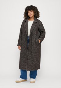 Lang, brun herringbone frakk med slagkrage, knappelukking og sidelommer, båret over en hvit topp og blå jeans.