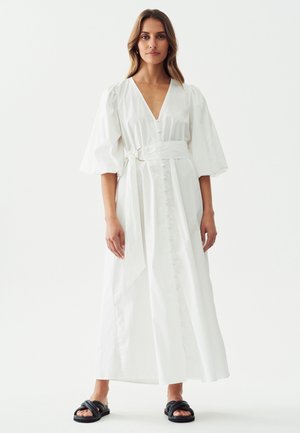 Vestido de algodón blanco con un escote en V profundo y mangas abullonadas, que presenta un frente con botones y un lazo en la cintura. Sandalias negras completan el conjunto.
