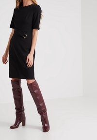 Svart klänning med korta ärmar, åtsittande midja och guld cirkulär detalj. Burgunder knähöga stövlar med en glänsande lädertextur och kraftiga klackar.