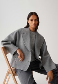 Vrouw zit op een houten stoel draagt een oversized grijze wollen jas, een grijze trui en donkergrijze broek tegen een effen lichte achtergrond.