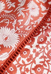 Motifs floraux rouges et blancs sur tissu, séparés par un galon tissé rouge texturé avec des dessins complexes de feuilles et de fleurs.