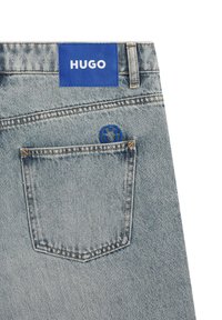 Jeansy denimowe o jasnym niebieskim odcieniu, z niebieską skórzaną łatą z napisem "HUGO" oraz małym haftowanym logo na tylnej kieszeni.