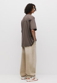 Smeđa oversized majica s kratkim rukavima, uparena s labavim bež trapericama. Outfit je upotpunjen bijelim i narančastim tenisicama.