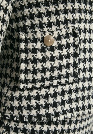 Tweedjasje met een zwart-wit houtsnip patroon, voorzien van een gouden knop en rafelranddetails. Gemêleerde stofafwerking.