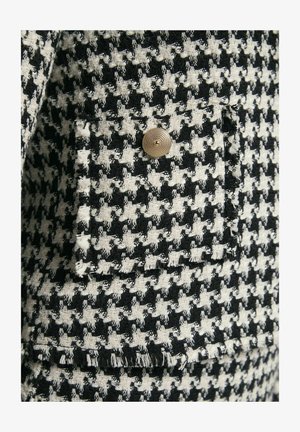 Tweedjasje met een zwart-wit houtsnip patroon, voorzien van een gouden knop en rafelranddetails. Gemêleerde stofafwerking.