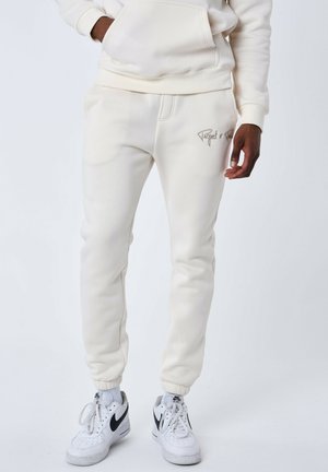 Sudadera y joggers de color crema en tejido suave. Cuenta con un bolsillo frontal, puños de canalé y un detalle de logotipo bordado. Combinado con zapatillas blancas.