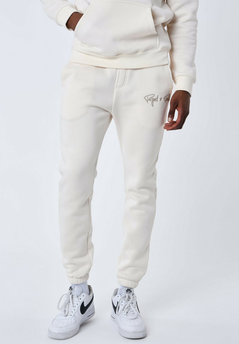 Felpa color crema e pantaloni jogger in morbido tessuto. Presenta una tasca frontale, polsini a coste e dettaglio del logo ricamato. Indossati con sneakers bianche.