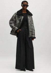 Grau und schwarze strukturierte Jacke mit einem großen schwarzen Pelzkragen, kombiniert mit weiten schwarzen Hosen. Sie hat Frontreißverschlüsse und Taschen.