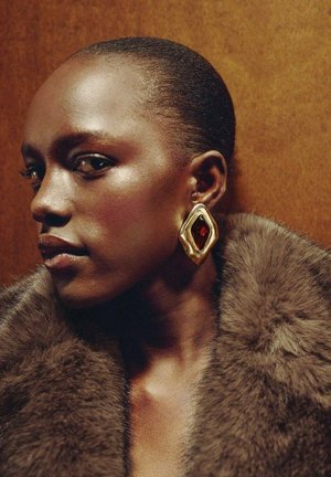 Goudkleurige statement oorbel met een diamante vorm, uitgerust met een centrale rode edelsteen, gecombineerd met een dikke, bruine bontkraag.