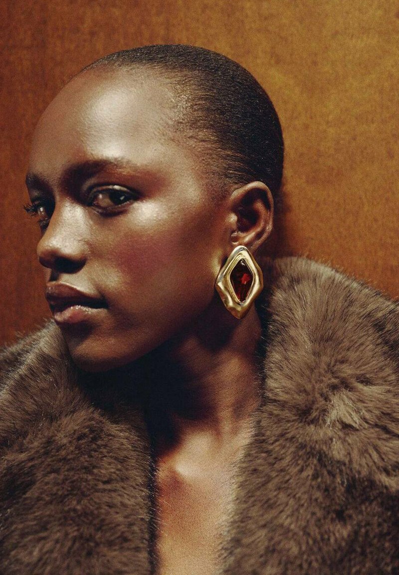 Komentárové náušnice zlatého odtieňa s diamantom v tvare, so stredným červeným drahokamom, kombinované s hrubým hnedým kožušinovým golierom.