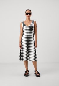 Robe midi rayée noir et blanc avec un décolleté en V et des boutons dorés sur le devant, portée avec des sandales noires. Texture lisse et côtelée.