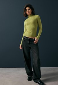 Gestreifter, langärmeliger Rollkragenpullover in leuchtendem Grün, kombiniert mit weit geschnittenen, hochtaillierten schwarzen Jeans und schwarzen Stiefeln. Flacher Hintergrund.