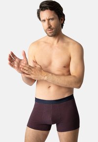 Boxer shorts à motifs rouge foncé et marine avec une taille élastique, présentant des designs géométriques et un ajustement serré. Le modèle les porte tout en gesticulant.