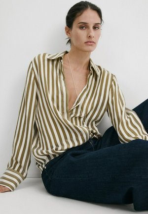 Femme assise contre un mur blanc, portant un chemisier à rayures verticales olive et blanc et un jean bleu foncé, avec une expression faciale neutre.