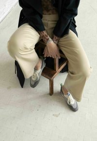 Pantaloni beige a gamba larga abbinati a scarpe lace-up grigie e bianche, con accenti blu e verdi. Persona seduta su uno sgabello di legno.