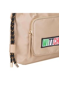 Sac en nylon beige avec un patch brodé bien en vue, étiqueté "WITTCH", et des accents contrastés noir, orange et blanc. Doté de poches zippées.