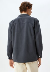 Camisa de manga larga gris oscuro, con cuello y puños abotonados. Textura suave, costuras visibles y un detalle de canesú en la parte posterior.