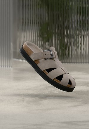 Beežist seemisnahast sandaal musta tallaga, korkjalanõu, reguleeritava pannaliga rihmaga ja mitme lõigatud rihmaga, hõljumas üle sileda betoonpõranda.