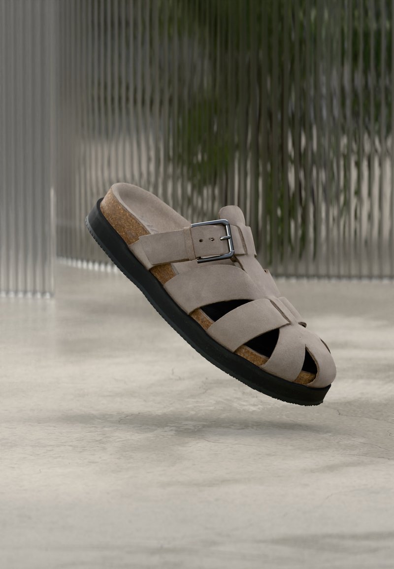 Sandale en suède beige avec semelle noire, semelle intérieure en liège, bride ajustable à boucle et plusieurs brides découpées, flottant au-dessus d'un sol en béton lisse.