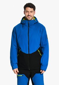 Blaue und schwarze isolierte Jacke mit Reißverschluss, Kapuze und grünen Akzenten. Entworfen mit einem geometrischen Farbblockmuster und seitlichen Taschen.